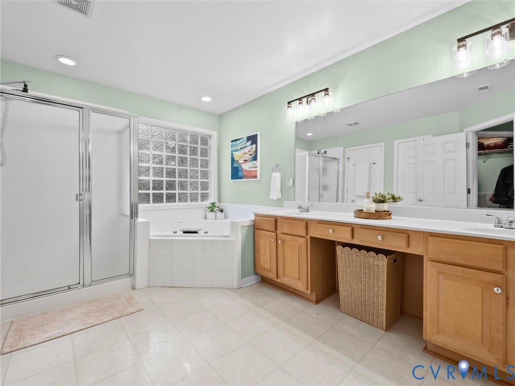Spacious bathroom featuring a glass-enclosed shower, a large soaking tub with tile surround, a dual vanity with light wood cabinetry, and a full-width mirror