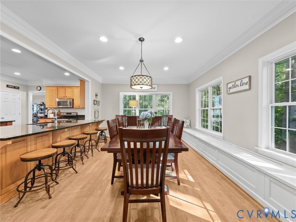 Open-concept dining area with light wood-finish flooring, recessed lighting, built-in window seating, and a large pass-through to the kitchen featuring a black countertop