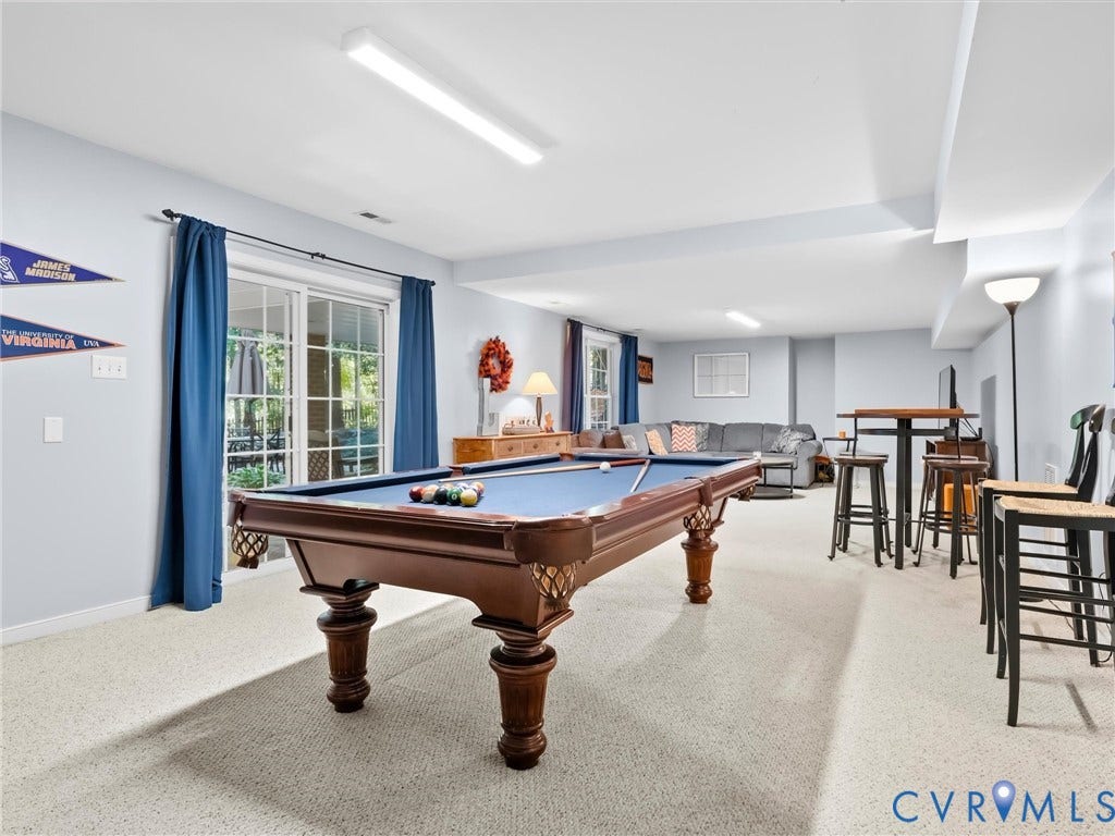 Spacious recreation room featuring a wood-finish billiards table with ornate turned legs, light-filled interior with recessed and linear ceiling lighting, wall-to-wall carpeting, and large multi-pane windows with blue drapery