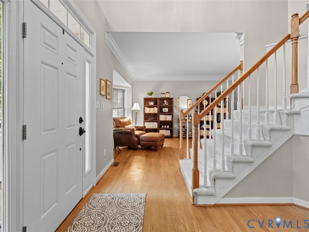Hardwood flooring throughout, white paneled door with transom window, carpeted staircase with wood handrail, and white balusters, and a crown molding detail