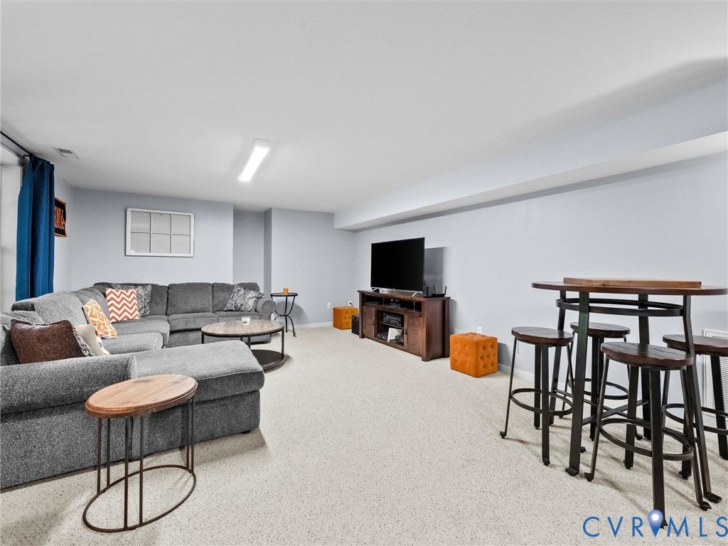 Spacious basement recreation area featuring neutral carpeting, recessed and linear ceiling lighting, a gray sectional sofa with coordinating side table, entertainment center with television, and a high-top dining counter with seating for four