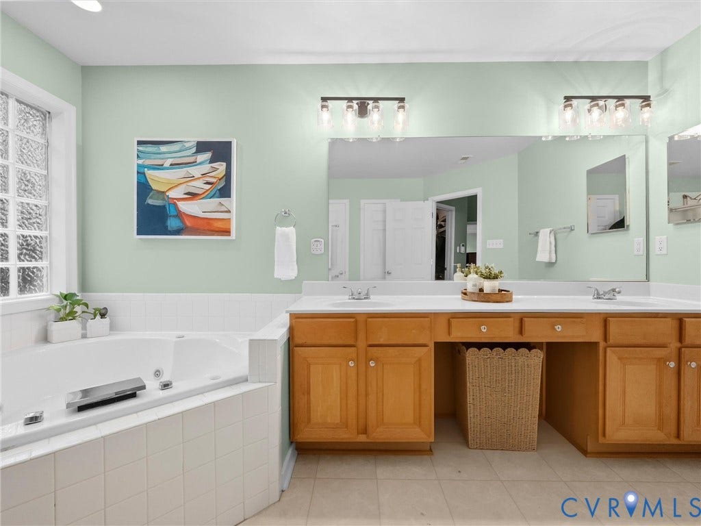 Dual vanity with wood cabinetry and white countertops, large soaking tub with tiled surround, and multiple wall-mounted light fixtures
