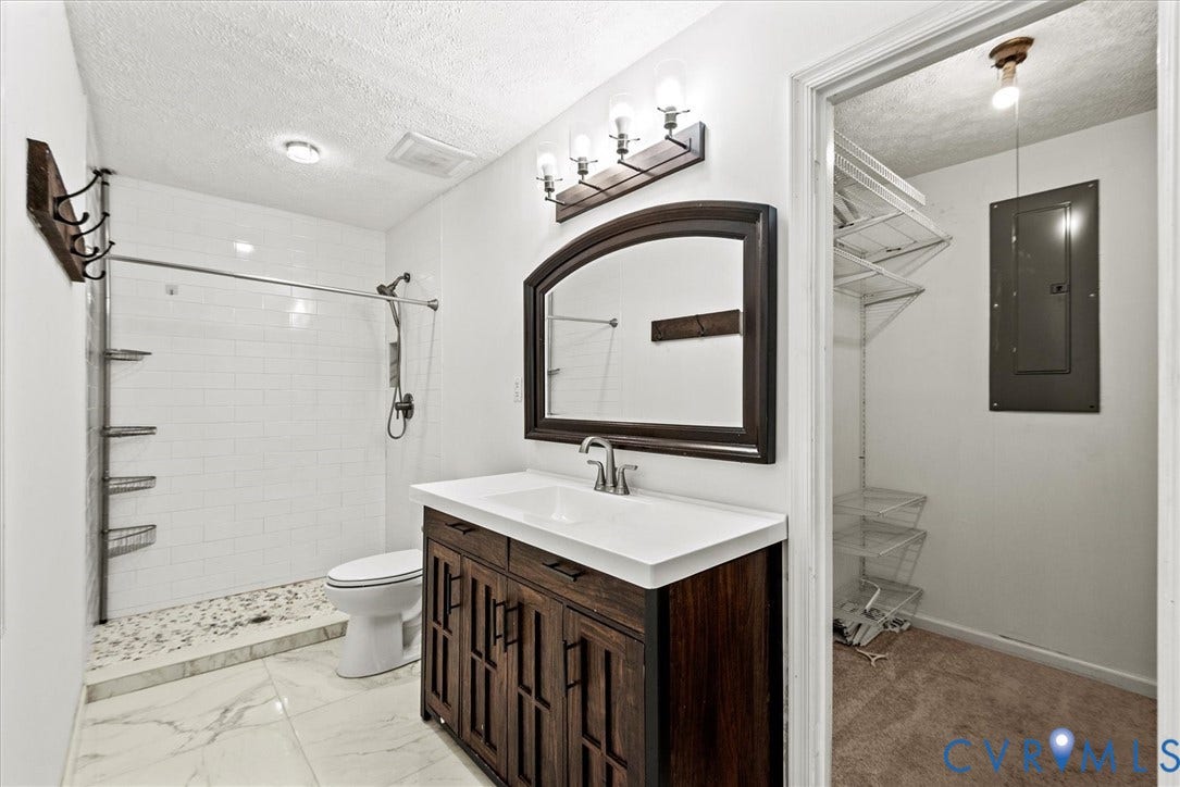 White subway tile shower with a river rock floor, dark wood-finish vanity with a white countertop and rectangular undermount sink, framed arched mirror, and white marble-look floor tile