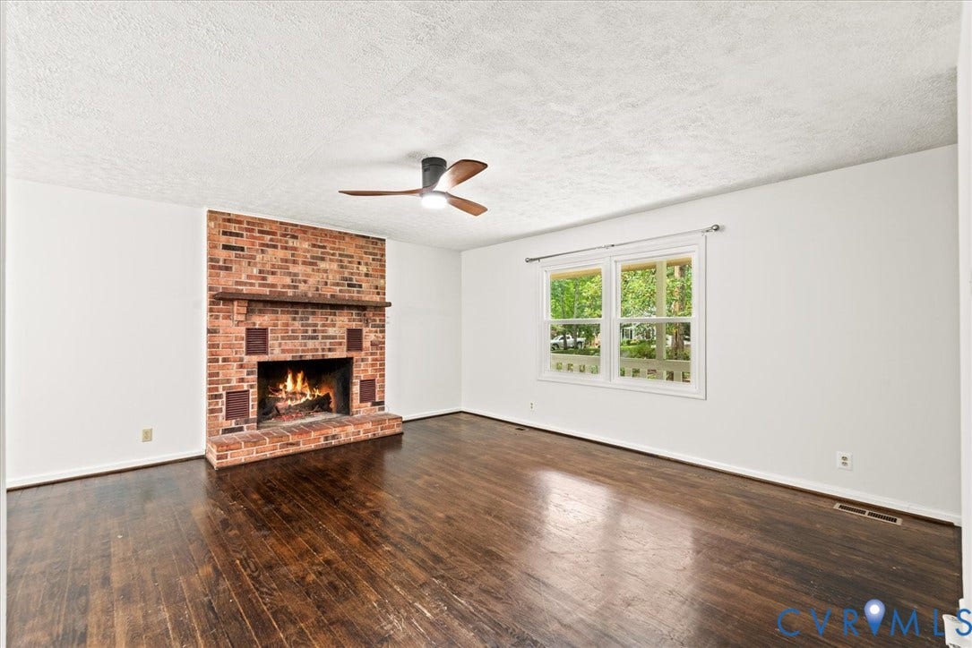 Exposed brick fireplace with a wood mantle, wood-finish flooring, white walls, and a ceiling fan