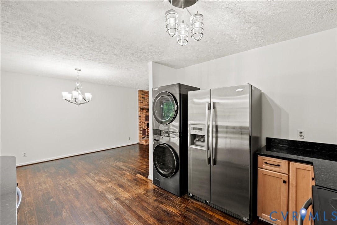 Stainless steel refrigerator, stacked washer and dryer, wood-finish flooring, dark countertops, and overhead lighting fixtures