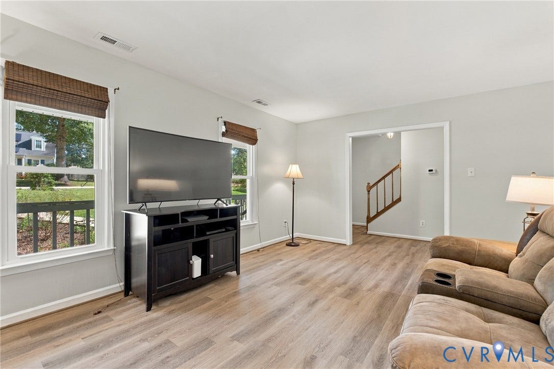 Neutral-toned living space featuring wood-finish flooring, dual windows with woven shades, and a wooden staircase railing