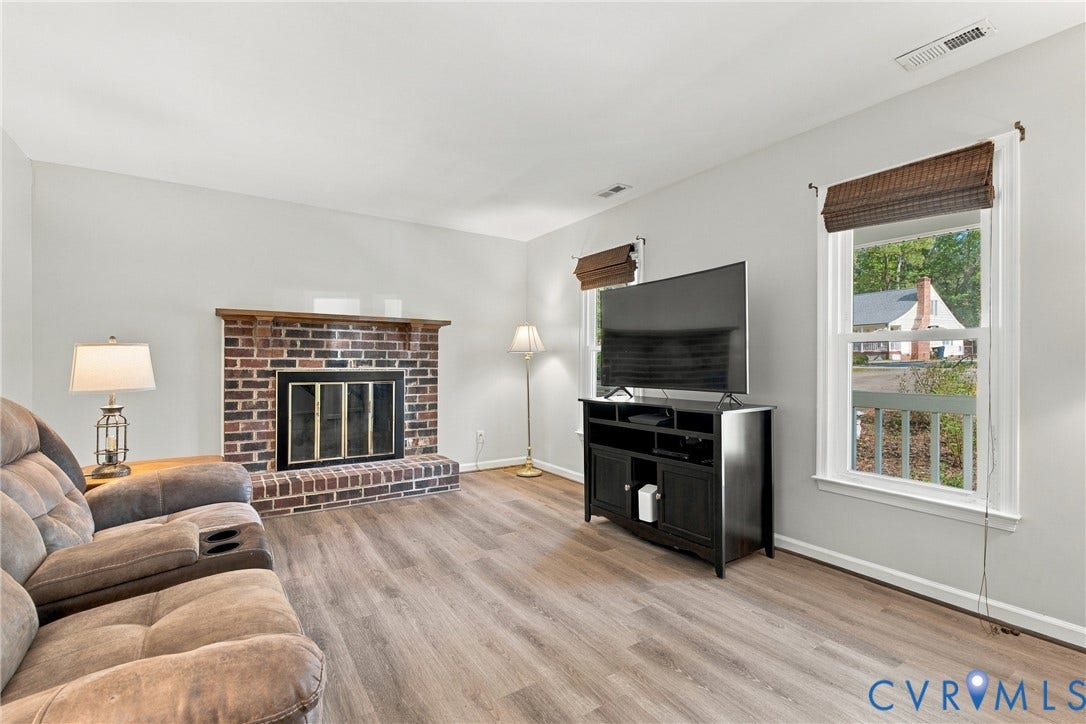 Brick fireplace with a wood mantel, wood-finish flooring, and multiple windows with integrated window treatments