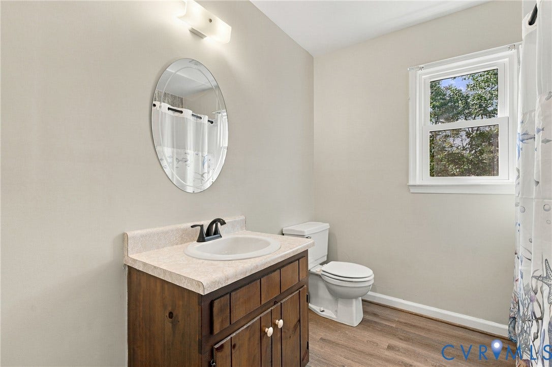 Bathroom featuring wood-finish flooring, a vanity with a stone-composite countertop and integrated sink, a wall-mounted oval mirror, and an exterior window
