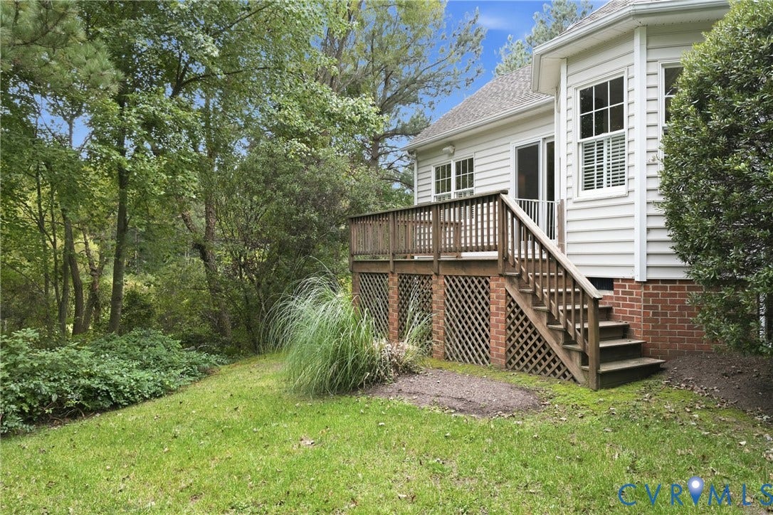 Spacious wooden deck with stairs, white siding, brick foundation, and mature trees surrounding a green lawn