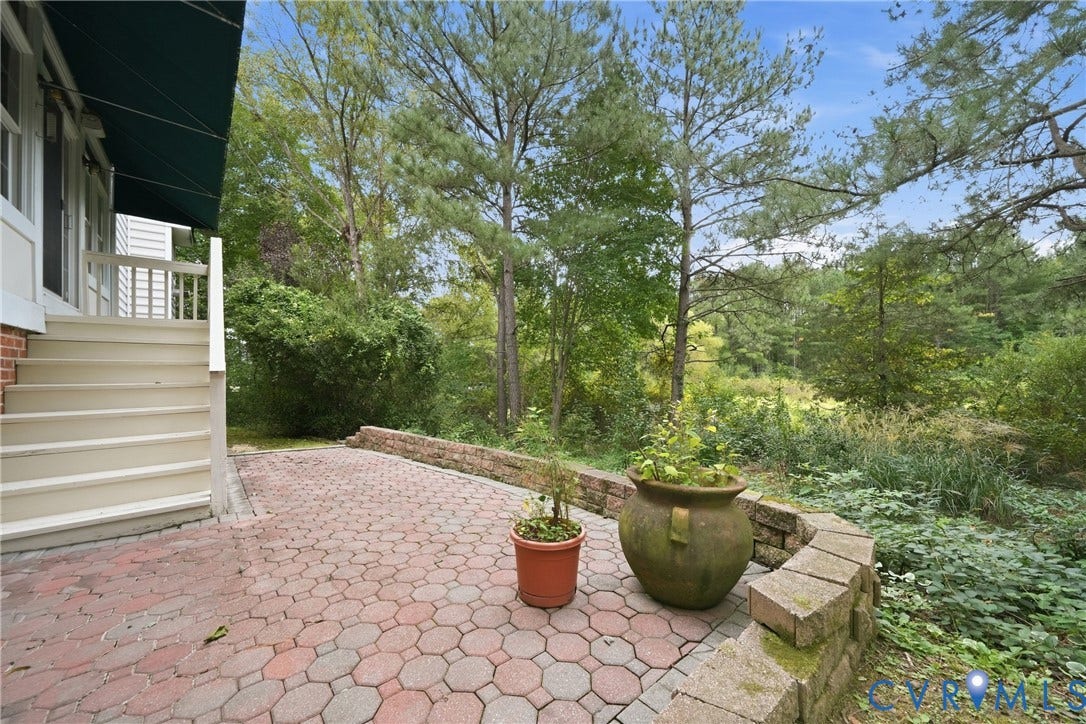 Brick paver patio with a stone retaining wall, wooden stairs, and dense natural landscaping