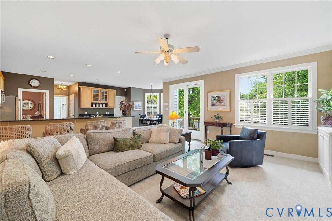 Open-concept living area with carpet flooring, a ceiling fan, and multiple windows with plantation shutters