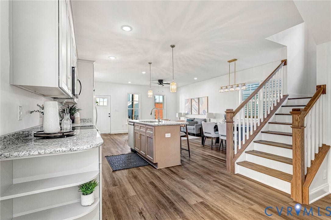 Open-concept living space featuring wood-finish flooring, a kitchen island with seating, a staircase with wood newel posts and handrail, recessed lighting, and pendant light fixtures