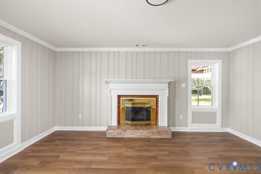 Living area with wood-finish flooring, a white mantel fireplace with a brick hearth, and two windows featuring white trim