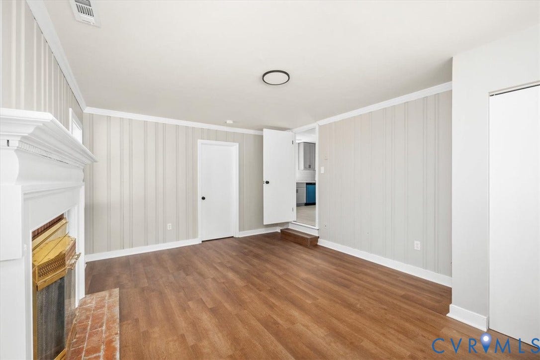 Spacious room featuring wood-finish flooring, vertical panel wall accents, a decorative fireplace with a brick hearth, and bright white trim