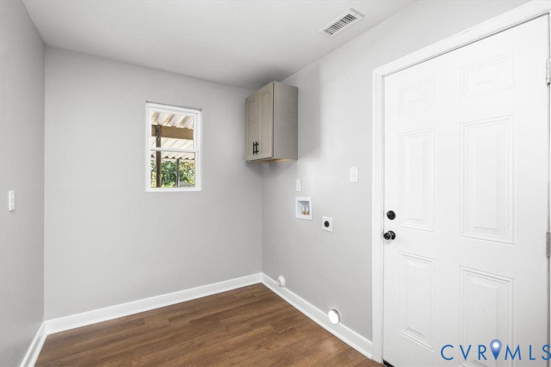 Wood-finish flooring, light gray walls, built-in wall cabinet, panel door with black hardware, and a single window