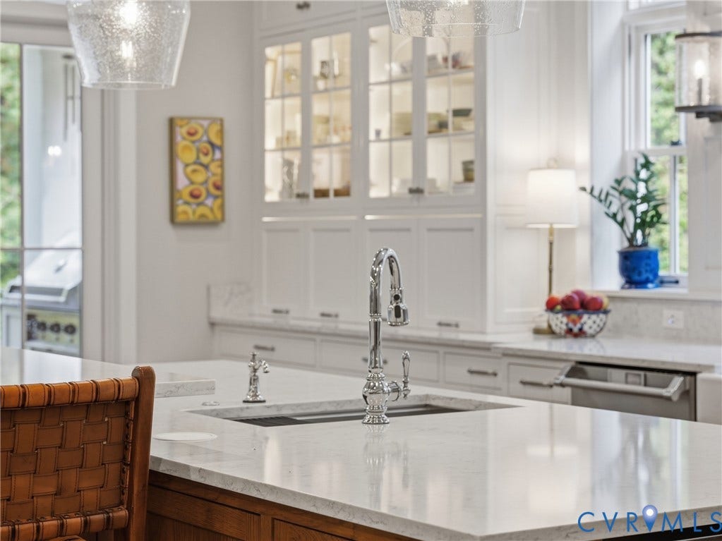 Kitchen featuring white cabinetry with glass-front display uppers, expansive stone-surface island, undermount sink with polished chrome bridge faucet, and stainless steel integrated dishwasher