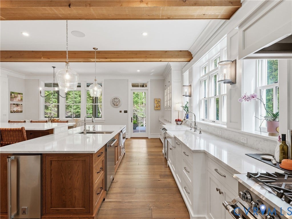 White cabinetry with ample storage, professional-grade stainless steel appliances, wood-finish flooring, exposed ceiling beams, and a large center island with integrated sink