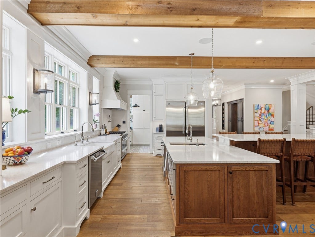 Gourmet kitchen featuring a large central island, light wood-finish flooring, white cabinetry, stainless steel appliances, and exposed wood beams