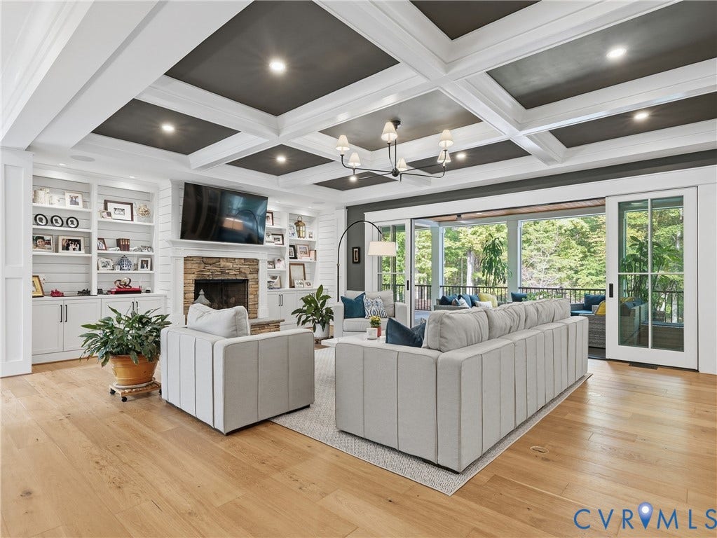 Coffered ceiling with recessed lighting and chandelier, stone-surround fireplace, built-in shelving, wood-finish flooring, and sliding glass doors to outdoor area