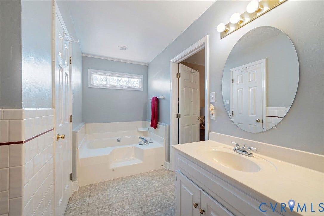 717 McCauliff Drive, Chesterfield, VA 23236 Soaking tub with a tiled surround, double vanity with a white countertop and basin sink, and an oval mirror with a gold-tone light fixture