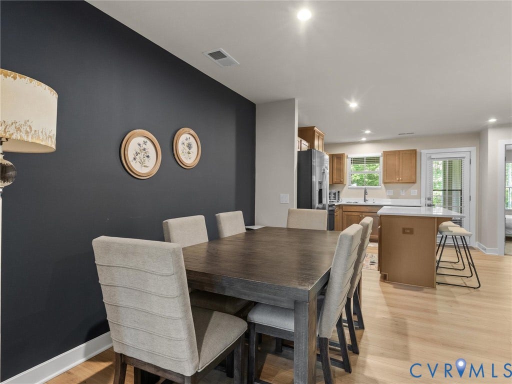 Light wood-finish flooring extending into the kitchen, kitchen island with counter seating, stainless steel refrigerator, light wood cabinetry, and recessed ceiling lighting