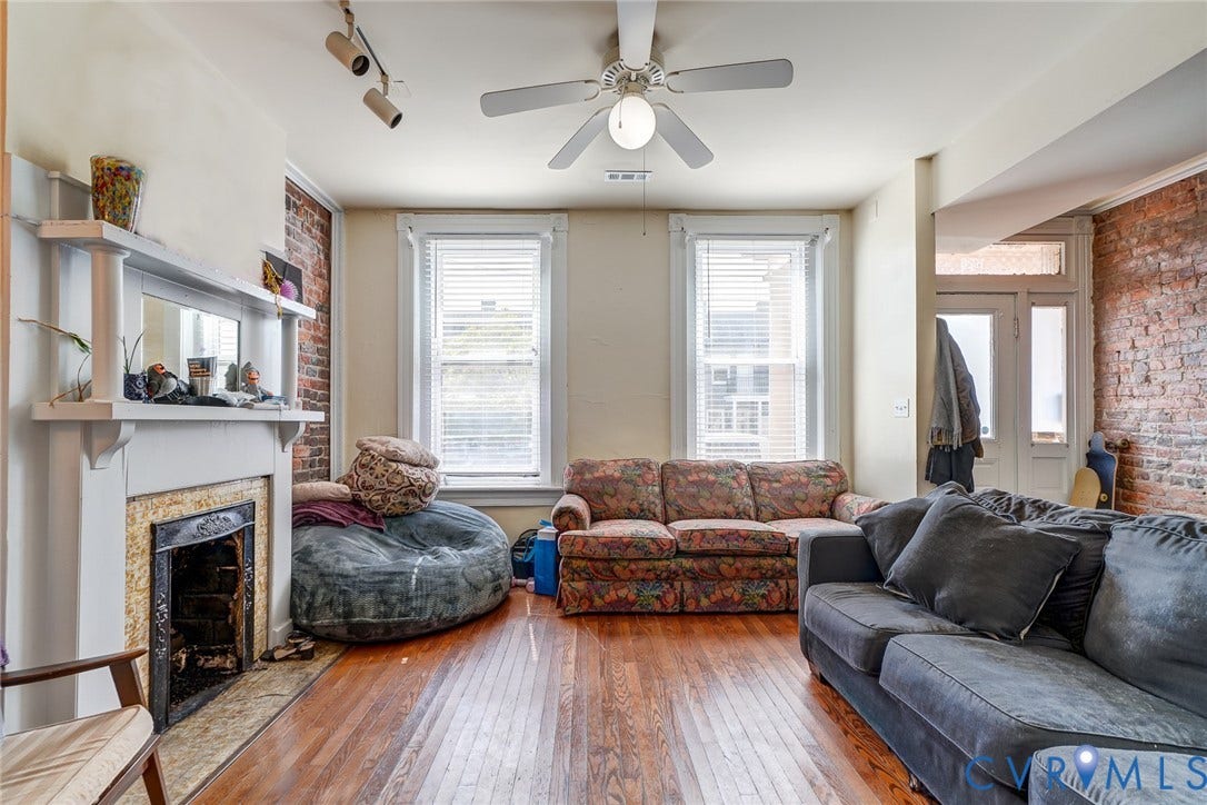 1620 W Cary Street, Fan Extenstion, Richmond, VA 23220 Hardwood flooring, exposed brick wall, white fireplace surround, two windows with blinds, and a ceiling fan with integrated lighting