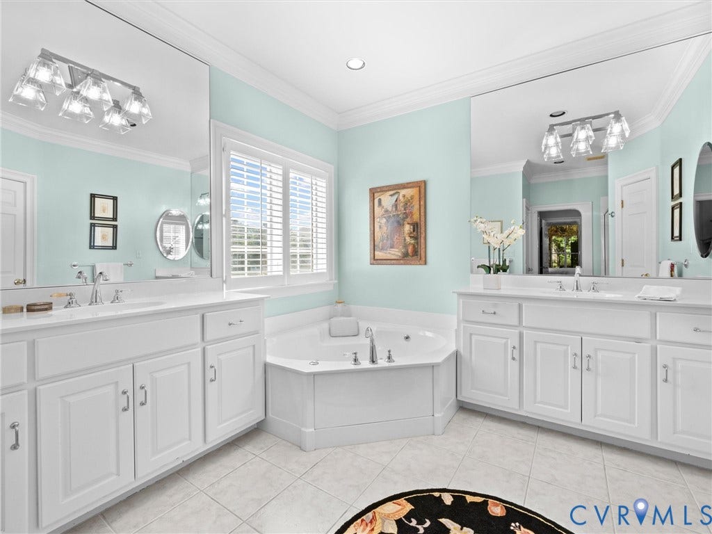 Dual vanities with white cabinetry and integrated sinks, corner built-in soaking tub, window with plantation shutters, and large mirror
