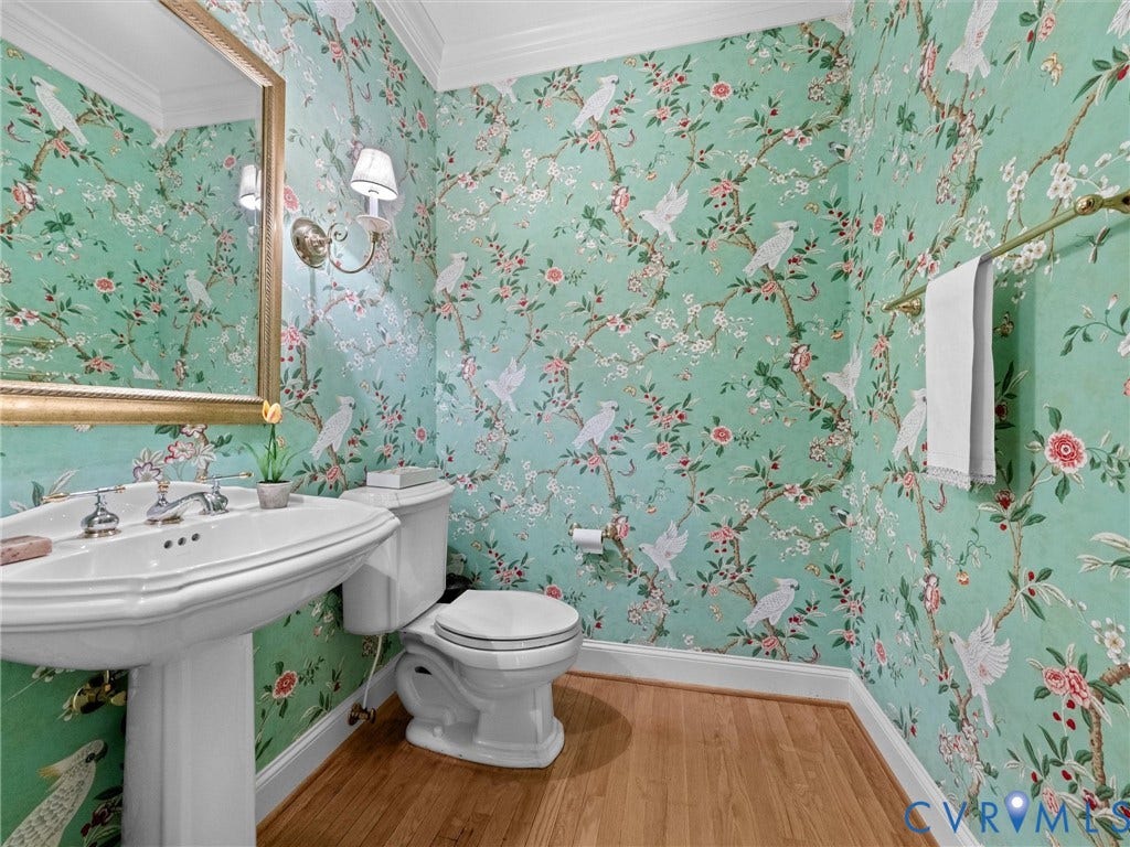Pedestal sink with chrome fixtures, gold-framed mirror, decorative wall sconce, wood-finish flooring, and crown molding