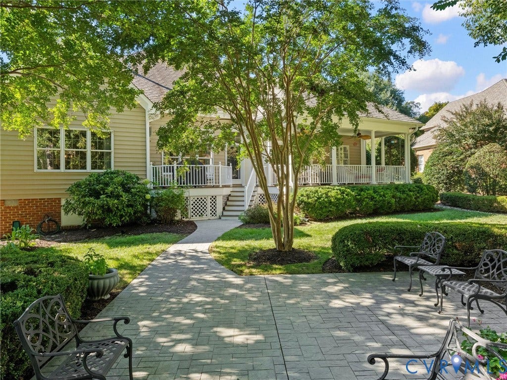 Expansive patio with pavers, manicured lawn, established landscaping, covered porch with white railings, and a side entrance with stairs