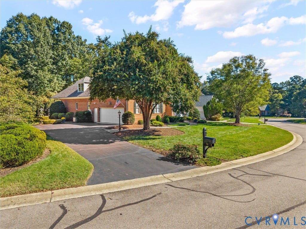 Brick exterior with white trim, attached garage, dormer window, paved driveway, and mature landscaping