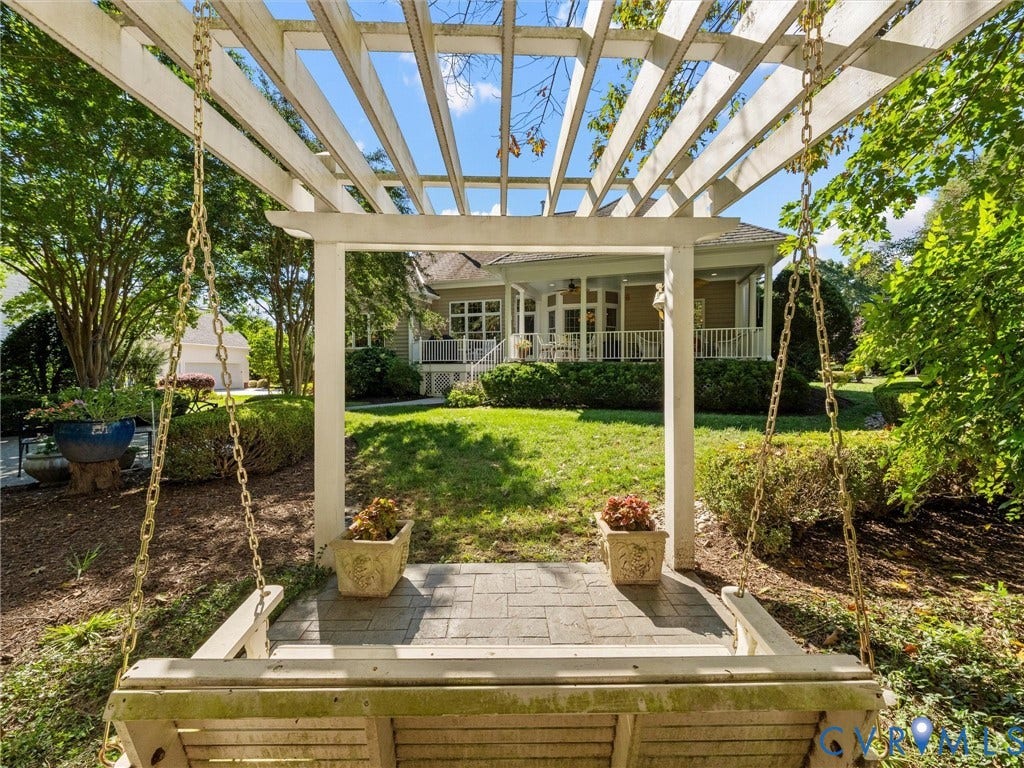 Pergola with a porch swing overlooking a manicured lawn, framed by lush landscaping and a distant covered porch with white railings