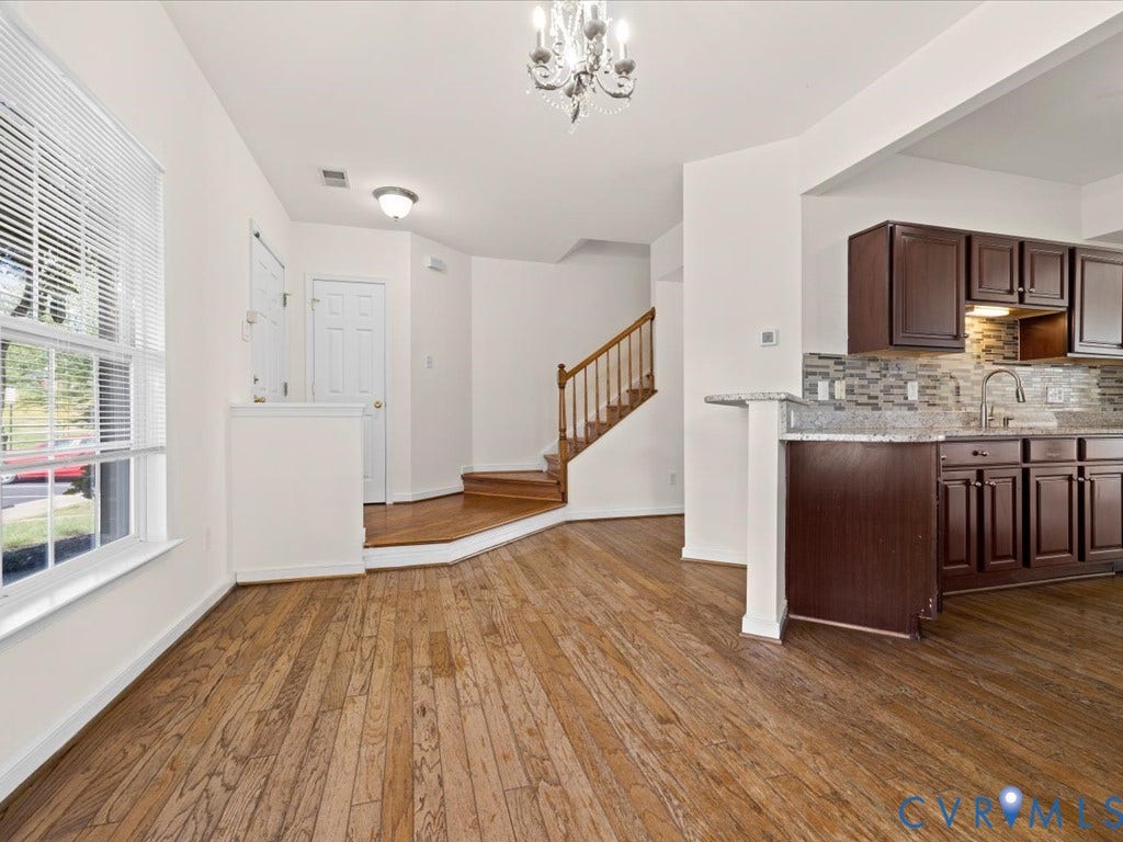 Wood-finish flooring throughout, staircase with wood handrail, kitchen with dark wood cabinetry and stone countertops, tile backsplash, and a decorative chandelier