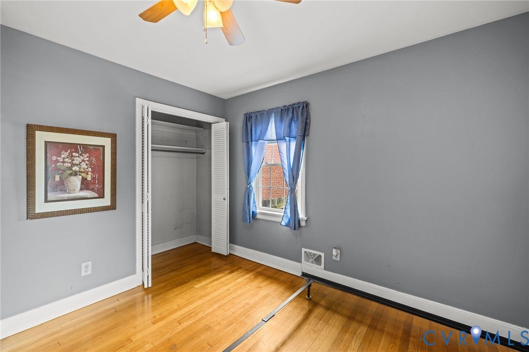 2405 Dumbarton Road, Henrico, VA 23228 Spacious bedroom with warm wood-finish flooring, neutral gray walls, and abundant natural light from a street-facing window with blue curtains