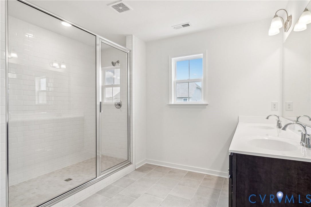 9116 TERROIR Lane, Viniterra Villas, New Kent, VA 23124 Dual vanity with white countertop and integrated sinks, glass-enclosed shower with white subway tile, tiled flooring, and a window providing natural light
