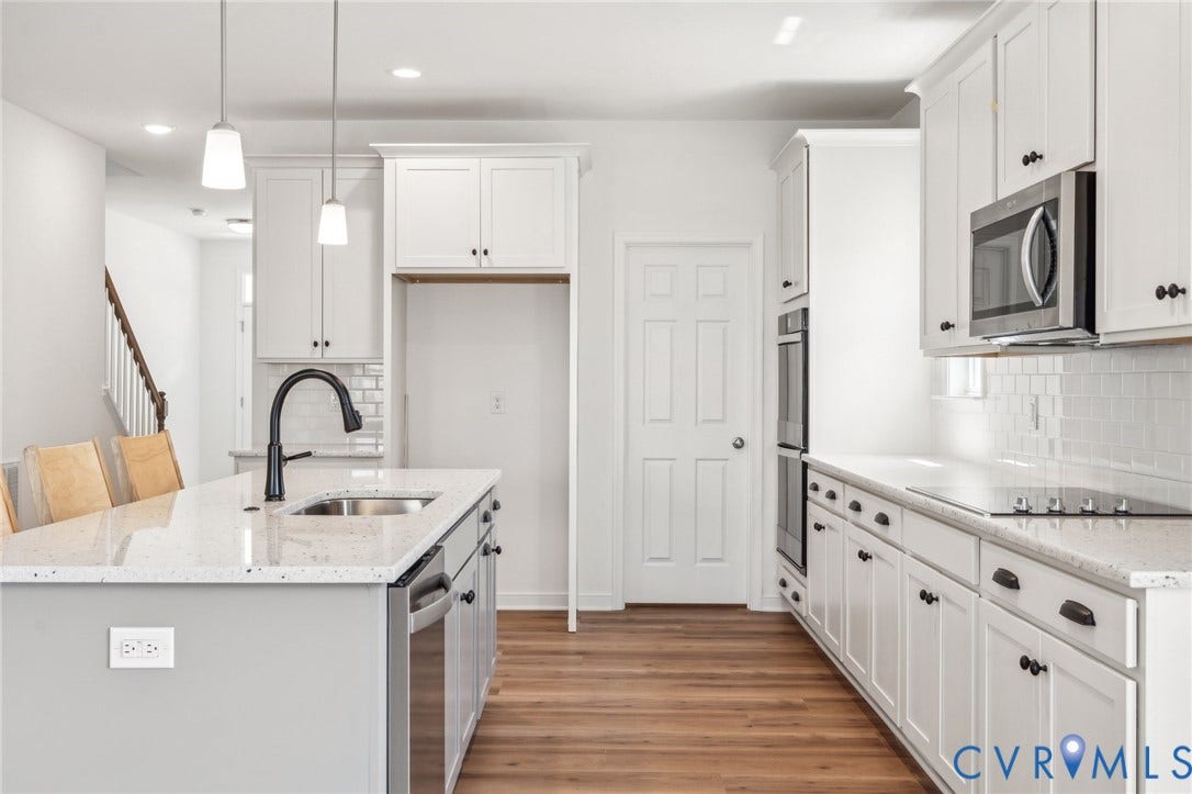9116 TERROIR Lane, Viniterra Villas, New Kent, VA 23124 Well-appointed kitchen featuring an island with an undermount sink, pendant lighting, built-in stainless steel appliances, white cabinetry, and wood-finish flooring