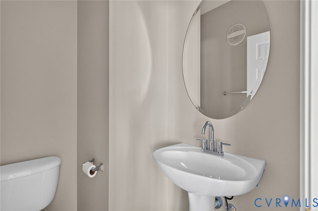 Modern powder room featuring a sleek oval mirror with polished chrome frame, a curved pedestal sink with chrome faucet, neutral taupe wall finishes, and ceramic tile flooring