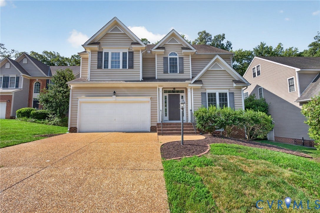 Two-story residence with a light-colored siding exterior, an attached garage, a textured concrete driveway, and established landscaping