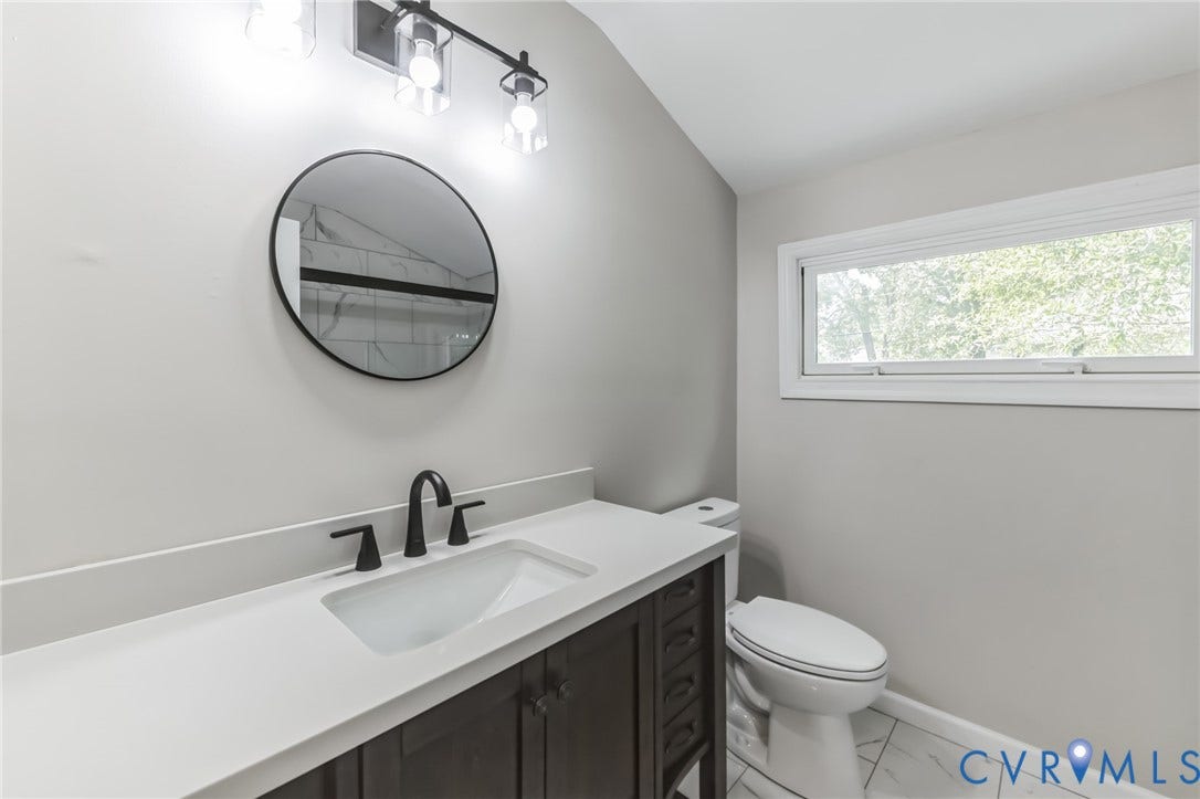 White countertop vanity with integrated sink and matte black faucet, dark wood-finish cabinetry, round mirror, and tile flooring