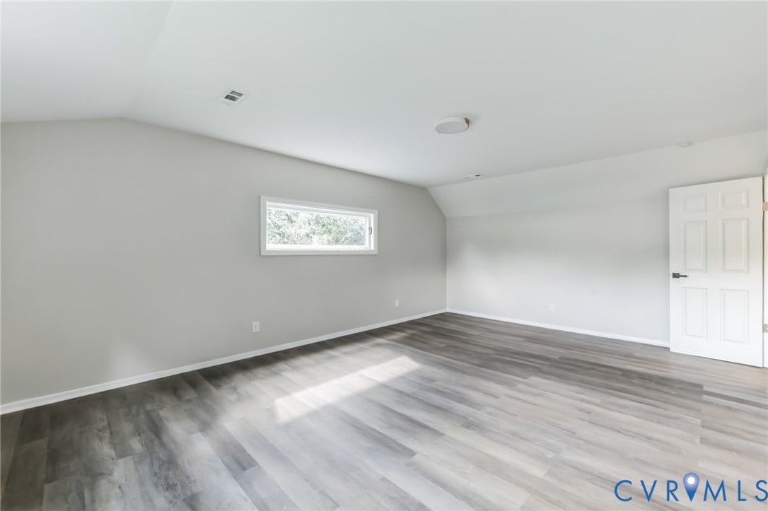 Expansive room featuring wood-finish flooring, a window with white trim, a vaulted ceiling, and a white paneled door