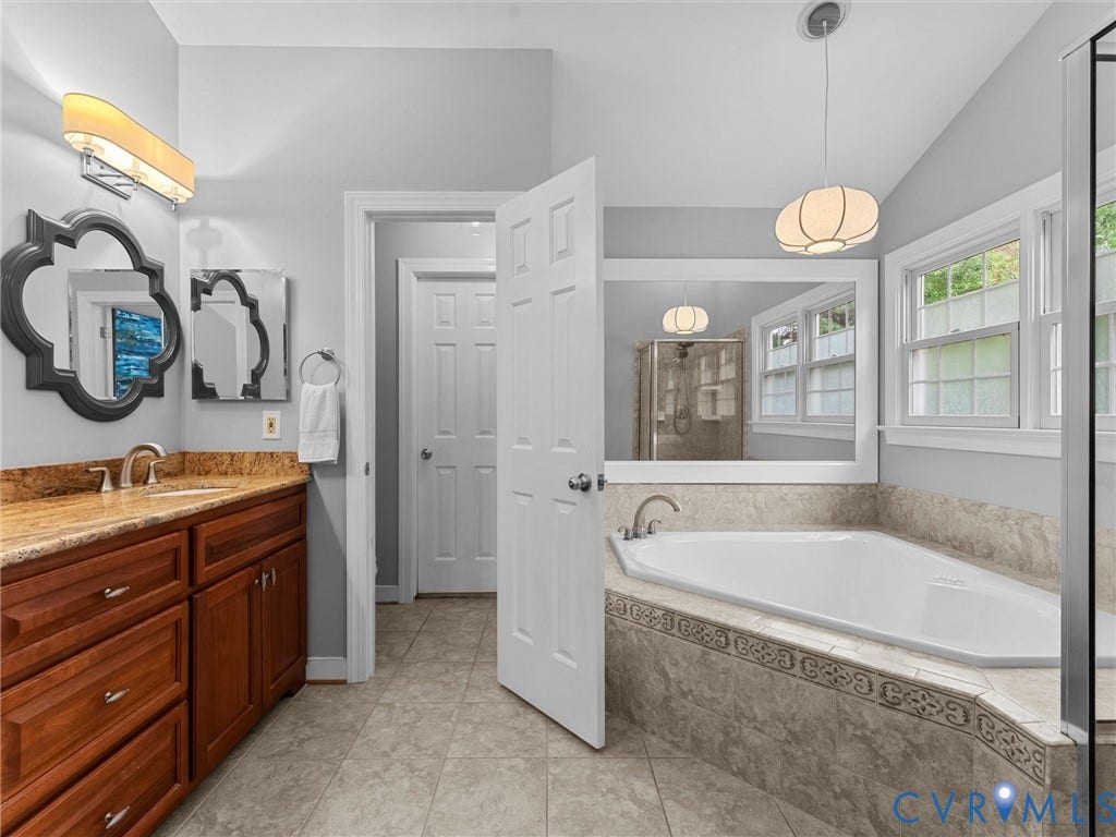 12100 English Ivy Lane, Glen Allen, VA 23059 Corner soaking tub with tile surround, wood-finish vanity cabinetry, stone countertop, decorative wall mirror, and pendant lighting
