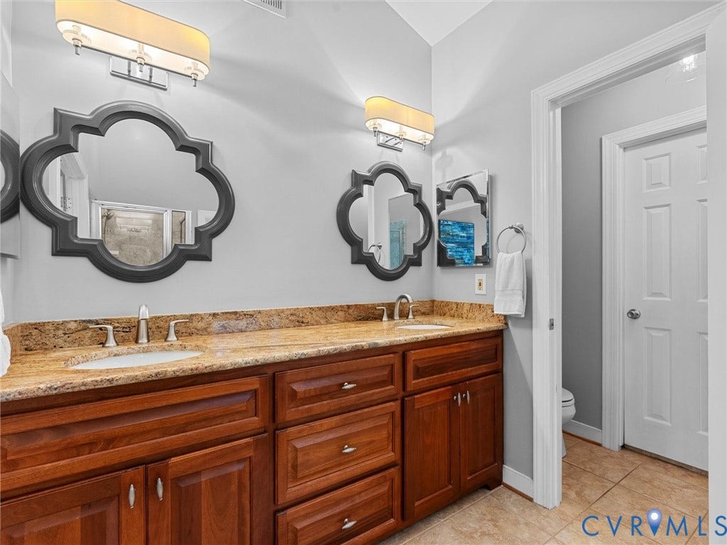12100 English Ivy Lane, Glen Allen, VA 23059 Double vanity featuring wood cabinetry, stone-finish countertops, dual drop-in sinks, decorative wall mirrors, and wall-mounted light fixtures