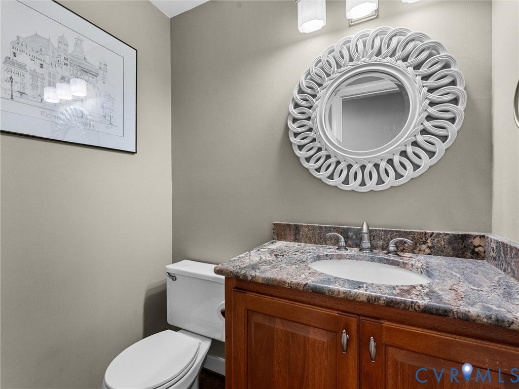 12100 English Ivy Lane, Glen Allen, VA 23059 Vanity featuring a natural stone countertop, undermount sink, and wood cabinetry, complemented by a round decorative wall mirror and multi-bulb vanity lighting