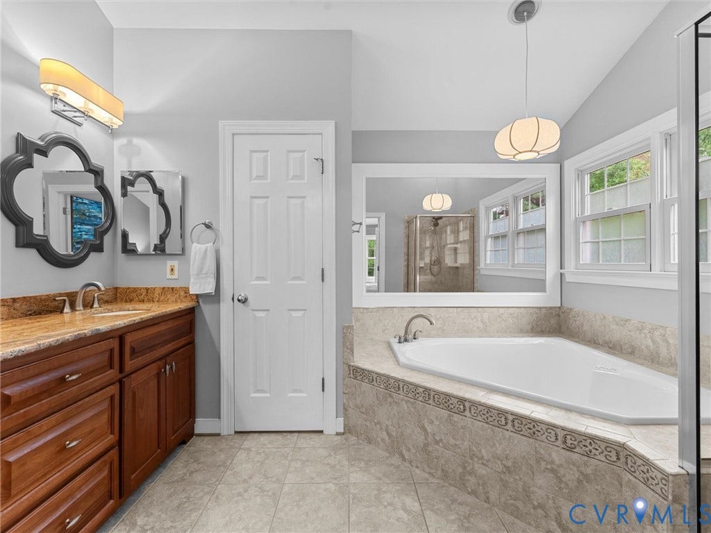 12100 English Ivy Lane, Glen Allen, VA 23059 Bathroom featuring a wood vanity with a stone countertop, a drop-in corner soaking tub with a tiled surround, a large wall-mounted mirror, and decorative light fixtures