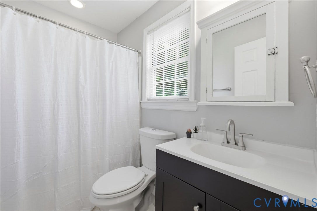 3015 M Street, Church Hill, Richmond, VA 23223 White shower curtain with metal rod, toilet, window with blinds, white medicine cabinet, and dark wood-finish vanity with white countertop and integrated sink