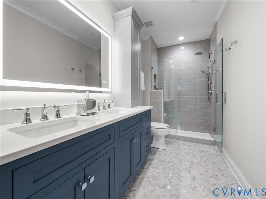 511 Libbie Avenue, #A, The Row At Westhampton, Richmond, VA 23226 Dual vanity with white undermount sinks and chrome fixtures, illuminated mirror, walk-in shower with subway tile surround, clear glass enclosure, and herringbone tile flooring