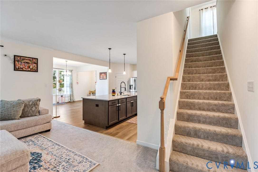 Carpeted staircase with wooden handrail, open concept living area with wood-finish flooring, kitchen island with dark cabinetry, and pendant lighting