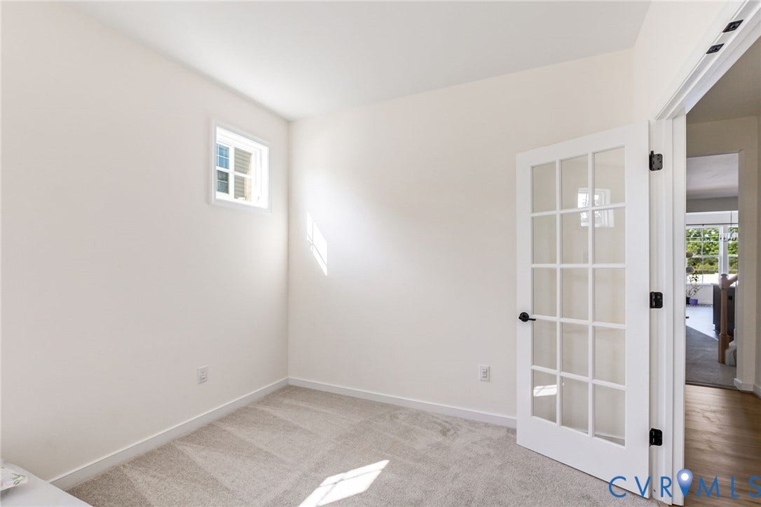 Neutral wall paint, carpet flooring, window with white trim, white French door with black hardware, and wood-finish flooring visible beyond the doorway