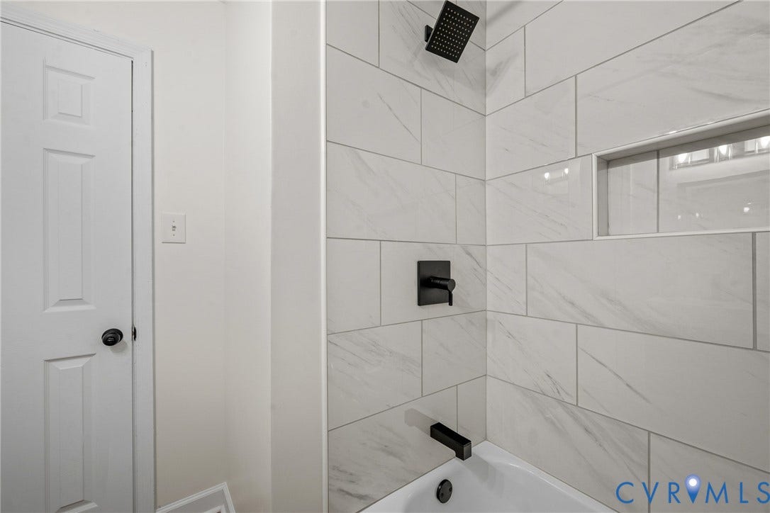 Bathtub with matte black fixtures, tiled surround featuring a recessed niche, and a contrasting white door