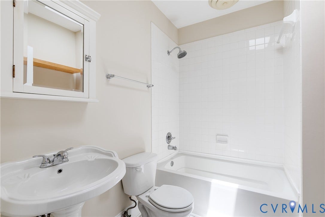 520 N 1st Street, Richmond, VA 23219 White pedestal sink with chrome faucet, white medicine cabinet with wood-finish shelving, white toilet, and white tiled shower/tub combination
