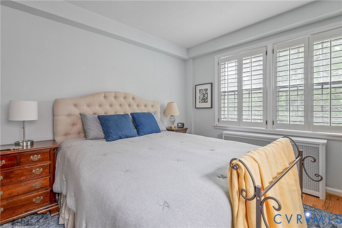 2606 Park Avenue, #U6, The Fan, Richmond, VA 23220 Tufted upholstered headboard, three windows with plantation shutters, wood-finish flooring, and a white radiator cover